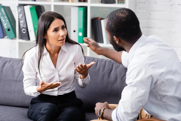 Selektiver Fokus eines afrikanisch-amerikanischen Psychologen, der frustrierte Patienten beruhigt, die auf dem Sofa sitzen und reden und gestikulieren — Stockfoto