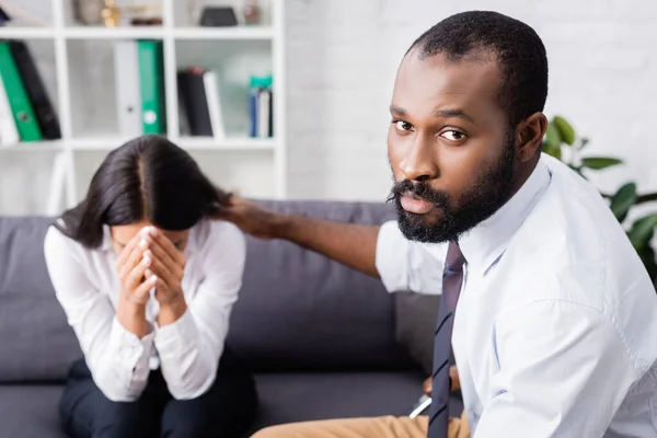 Serious african american psychologist looking at camera while calming crying woman obscuring face with hands — Stock Photo
