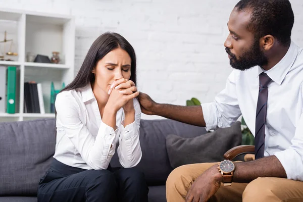 Psicólogo afroamericano calmar mujer molesta sentado en sofá con los ojos cerrados y las manos apretadas - foto de stock