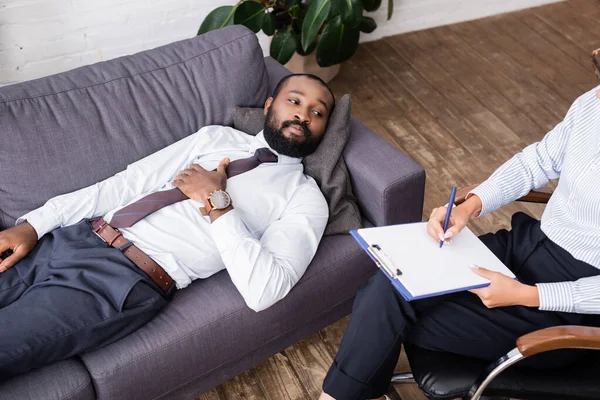 Vista ad alto angolo dell'uomo afro-americano in abbigliamento formale sdraiato sul divano vicino allo psicologo che scrive sugli appunti — Foto stock