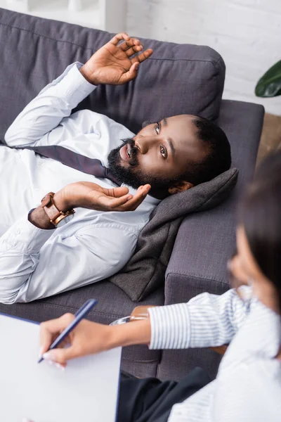 Selektiver Fokus des Psychologen, der auf Klemmbrett in der Nähe eines afrikanisch-amerikanischen Patienten spricht und gestikuliert, während er auf dem Sofa liegt — Stockfoto