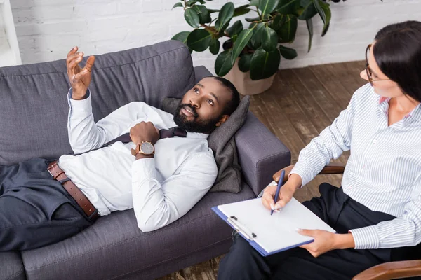 Enfoque selectivo del hombre afroamericano haciendo gestos y hablando mientras está acostado en el sofá cerca de psicólogo con pluma y portapapeles - foto de stock