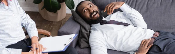 Imagen horizontal del hombre americano africano alegre que habla mientras que miente en el sofá cerca del psicólogo que sostiene el portapapeles vacío - foto de stock