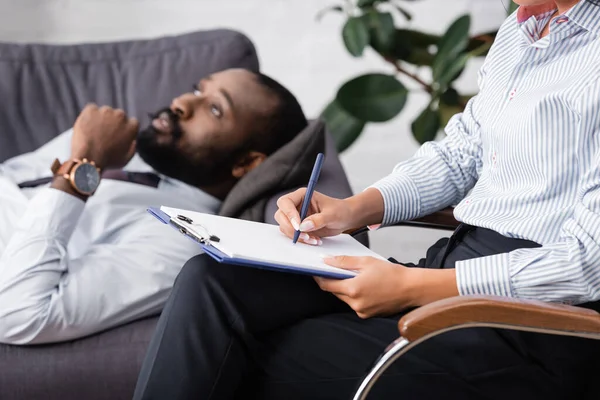 Selektiver Fokus eines nachdenklichen afrikanisch-amerikanischen Mannes, der auf dem Sofa in der Nähe eines Psychologen liegt und Rezepte auf Klemmbrett schreibt — Stockfoto