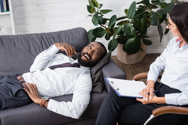 Homem americano africano alegre falando com psicólogo enquanto deitado no sofá — Fotografia de Stock