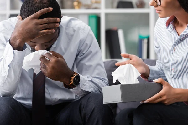 Psychologist holding paper napkins near stressed african american patient crying and touching head in hospital — Stock Photo