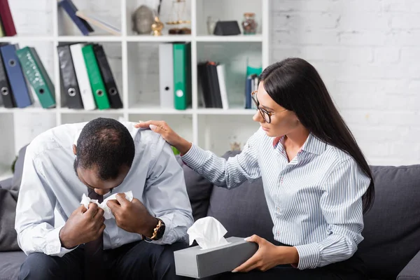 Morena psicólogo calmar africano americano hombre llorando y limpiando los ojos con papel servilleta mientras sentado en sofá - foto de stock