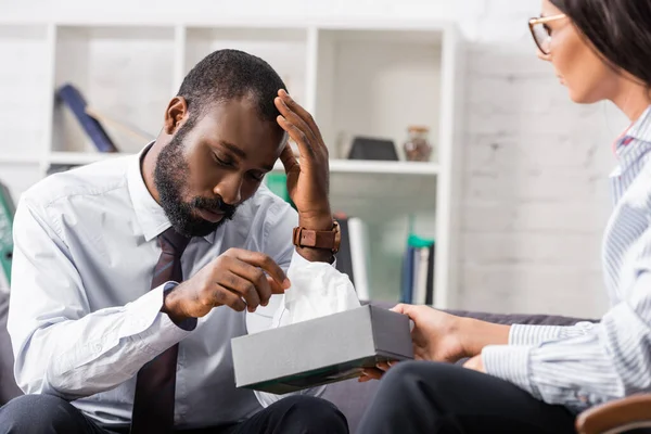 Foco seletivo de homem afro-americano frustrado tocando a cabeça enquanto toma guardanapo de papel do psicólogo — Fotografia de Stock