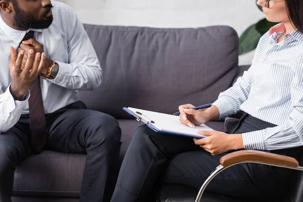 Vista ritagliata del paziente afroamericano gesticolare mentre parla con lo psicologo che tiene appunti e penna — Foto stock