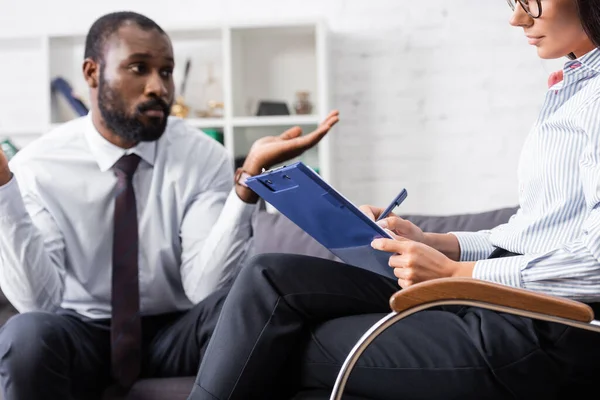 Selective focus of psychologist writing on clipboard near confused african american patient showing shrug gesture — Stock Photo