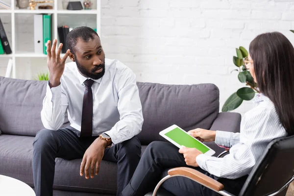 Selective focus of worried african american patient talking to brunette psychologist holding digital tablet with green screen — Stock Photo