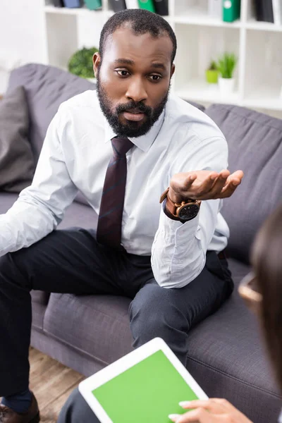 Enfoque selectivo del hombre afroamericano tenso gesto mientras habla con el psicólogo sosteniendo tableta digital con pantalla en blanco - foto de stock