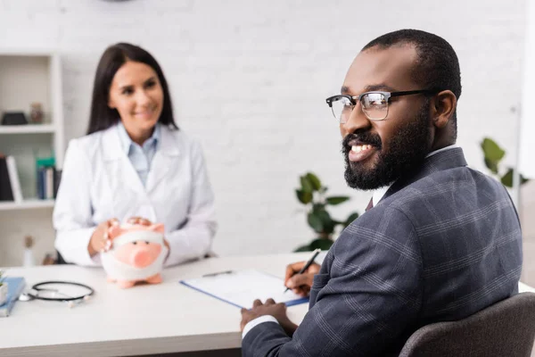 Joyeux homme afro-américain regardant la caméra près du médecin avec tirelire, enveloppé dans un bandage, concept d'assurance médicale — Photo de stock