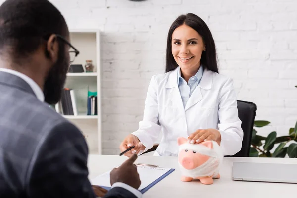 Brünette Ärztin berührt Sparschwein, in Verband gewickelt, und gibt Stift an afrikanisch-amerikanischen Patienten in der Nähe Versicherungsformular — Stockfoto