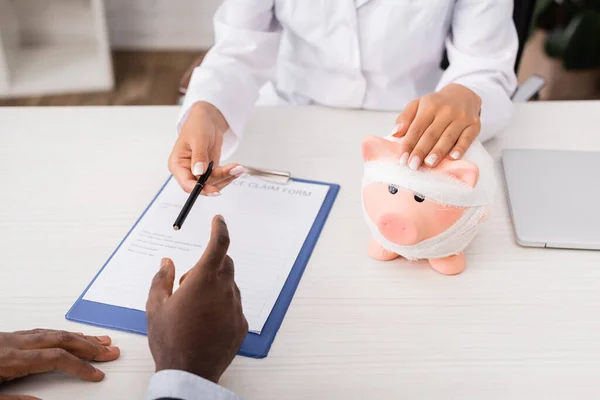 Vue recadrée du médecin touchant tirelire, enveloppé dans un bandage, et donnant stylo au patient afro-américain près du formulaire de demande de règlement d'assurance — Photo de stock