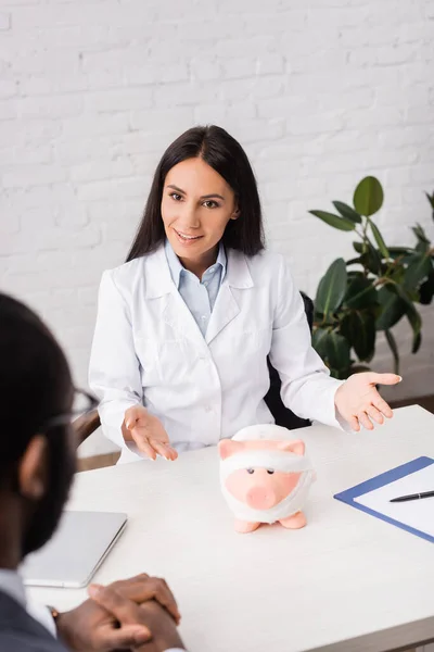 Selektiver Fokus des brünetten Arztes im Gespräch mit einem afrikanisch-amerikanischen Patienten in der Nähe eines Sparschweins, eingewickelt in einen Verband, Krankenversicherungskonzept — Stockfoto