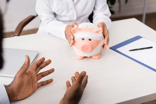 Cropped view of african american patient gesturing with hands near doctor touching piggy bank, wrapped in bandage, medical insurance concept — Stock Photo