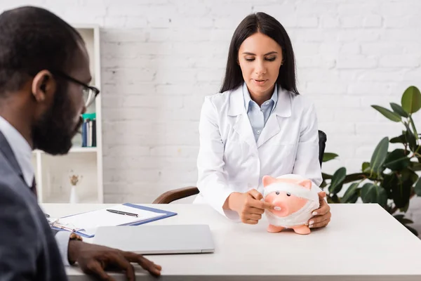 Brünette ärztin zeigt mit finger auf sparschwein, in verband gewickelt, in der nähe von afrikanisch-amerikanischem mann, krankenversicherungskonzept — Stockfoto