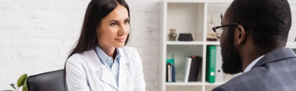 Panoramisches Konzept brünetter Arzt und afrikanisch-amerikanischer Patient, die sich im Krankenhaus anschauen — Stockfoto