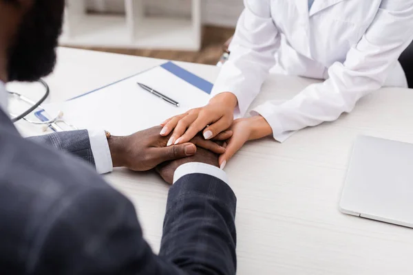 Visão parcial do médico tocando as mãos do paciente afro-americano perto clipboad no local de trabalho — Fotografia de Stock