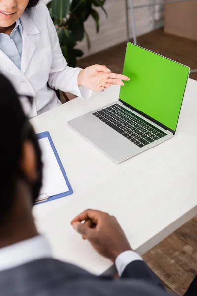 Vista recortada del médico apuntando con la mano a la computadora portátil con pantalla verde cerca de paciente afroamericano - foto de stock