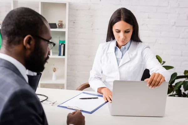 Enfoque selectivo de la computadora portátil de apertura de médico en el lugar de trabajo cerca de paciente afroamericano - foto de stock