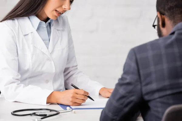 Vista recortada de la escritura del médico en el portapapeles cerca de paciente afroamericano - foto de stock