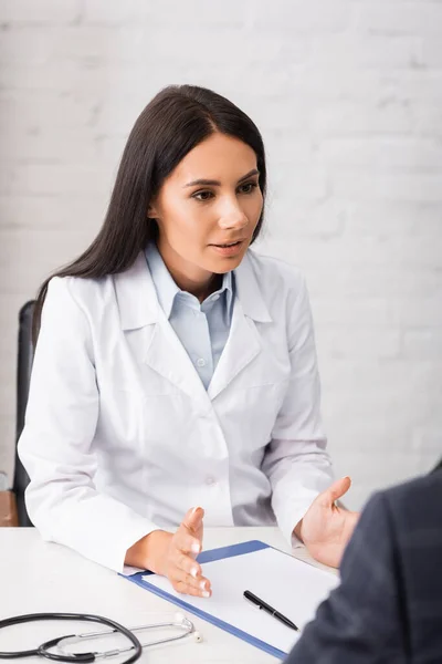 Selektiver Fokus des Arztes im Gespräch mit dem Patienten am Arbeitsplatz in der Nähe von Stethoskop und Klemmbrett — Stockfoto