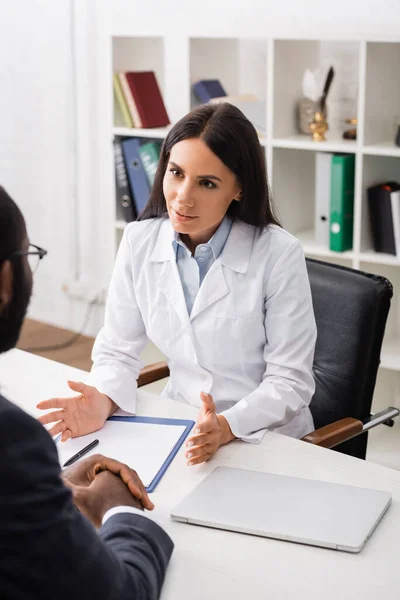 Brünette Ärztin gestikuliert im Gespräch mit afrikanisch-amerikanischer Patientin in der Nähe von Klemmbrett und Laptop — Stockfoto