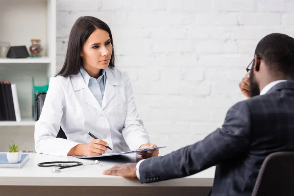 Selective focus of african american patient touching face while sitting opposite brunette doctor — Stock Photo