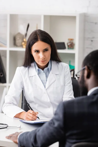 Selective focus of brunette doctor writing prescription on clipboard near african american patient — Stock Photo