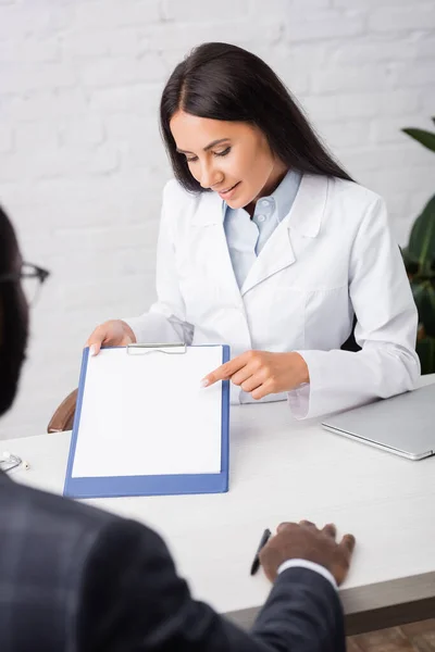 Morena médico apuntando con el dedo en el portapapeles con papel blanco cerca de hombre afroamericano - foto de stock