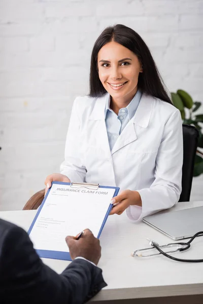 Brunette, joyeux médecin tenant presse-papiers tandis que le patient afro-américain signe formulaire de demande de règlement d'assurance — Photo de stock