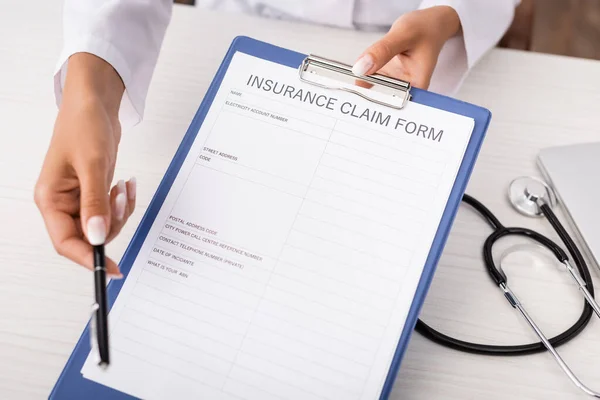 Partial view of doctor holding insurance claim form on clipboard near stethoscope on desk — Stock Photo