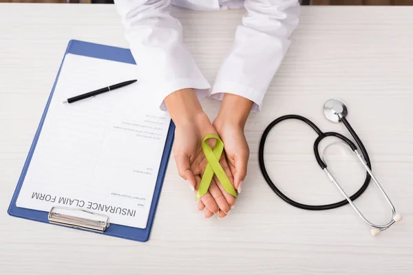 Vista superior del psicólogo que sostiene la cinta verde de la conciencia cerca del formulario de reclamación del seguro y del estetoscopio, concepto de salud mental - foto de stock