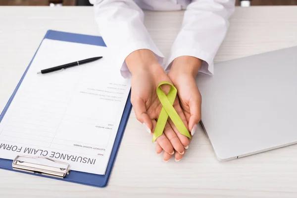 Vista recortada del psicólogo que sostiene la cinta verde de la conciencia cerca del formulario de reclamación del seguro y el ordenador portátil, concepto de salud mental - foto de stock