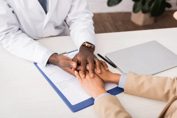 Teilansicht eines afrikanisch-amerikanischen Arztes, der die Hände eines Patienten in der Nähe von Versicherungsantragsformular und Laptop berührt — Stockfoto