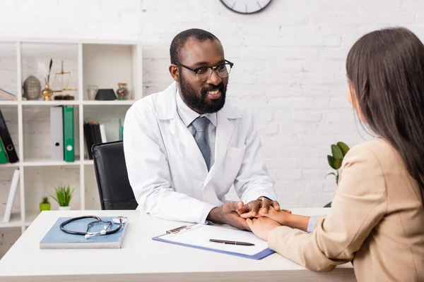 Afrikanisch-amerikanischer Arzt mit Brille berührt Hände eines brünetten Patienten in der Nähe von Klemmbrett mit Versicherungsformular — Stockfoto