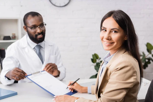 Selektiver Fokus der freudigen Frau, die in die Kamera schaut, während sie in der Nähe eines amerikanischen Arztes ein Versicherungsformular unterschreibt — Stockfoto