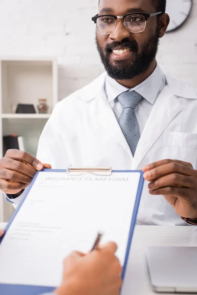 Selektiver Fokus des freudestrahlenden afrikanisch-amerikanischen Arztes, der Klemmbrett hält, während der Patient das Versicherungsantragsformular unterschreibt — Stockfoto