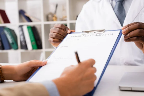 Vista ritagliata del paziente firma modulo di richiesta di assicurazione vicino afro-americano medico — Foto stock