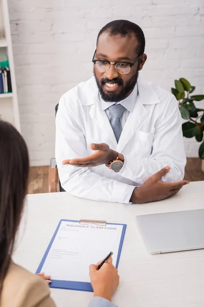 Foyer sélectif du médecin afro-américain dans les lunettes pointant avec la main près du patient signer formulaire de demande de règlement d'assurance — Photo de stock