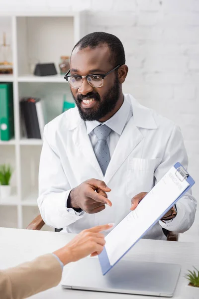 Selektiver Fokus von afrikanisch-amerikanischem Arzt und Patient, der mit dem Finger auf Klemmbrett mit Versicherungsformular zeigt — Stockfoto