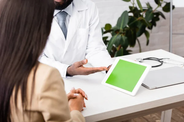 Ausgeschnittene Ansicht eines afrikanisch-amerikanischen Arztes, der mit der Hand auf ein digitales Tablet mit grünem Bildschirm in der Nähe einer brünetten Patientin zeigt — Stockfoto