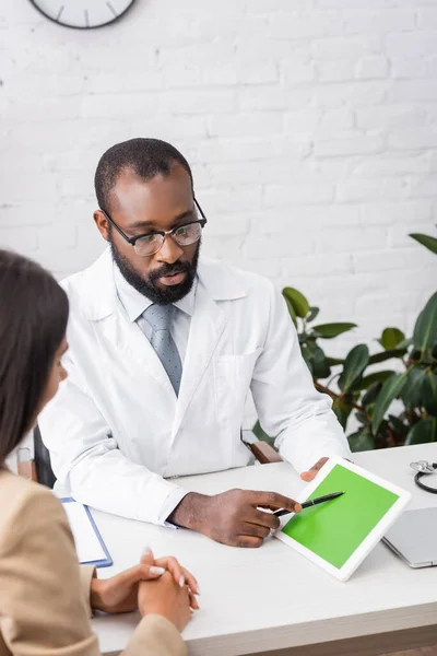 Foyer sélectif du médecin afro-américain grave dans les lunettes pointant avec stylo à tablette numérique avec écran vert près de la femme brune — Photo de stock