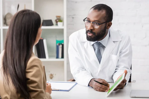 Selective focus of serious african american doctor showing digital tablet while talking to brunette woman — Stock Photo