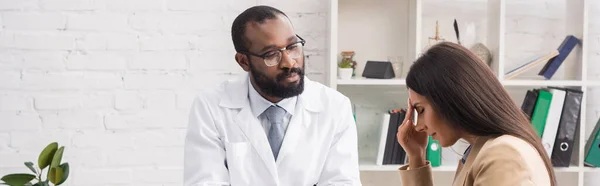 Colheita panorâmica de médico americano africano grave em óculos olhando para a mulher doente tocando a testa com os olhos fechados — Fotografia de Stock