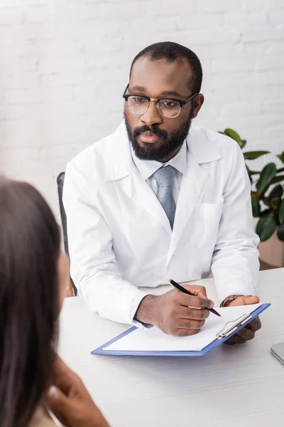 Africano americano médico em óculos segurando prancheta e caneta perto doente tocando dor de garganta — Fotografia de Stock