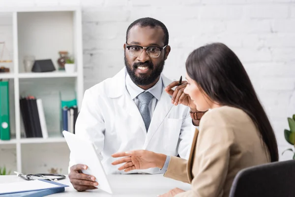 African american doctor holding digital tablet and looking at camera near brunette woman pointing with finger — Stock Photo