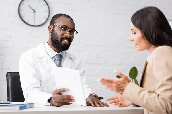 Selective focus of african american doctor in eyeglasses showing digital tablet to talking patient — Stock Photo